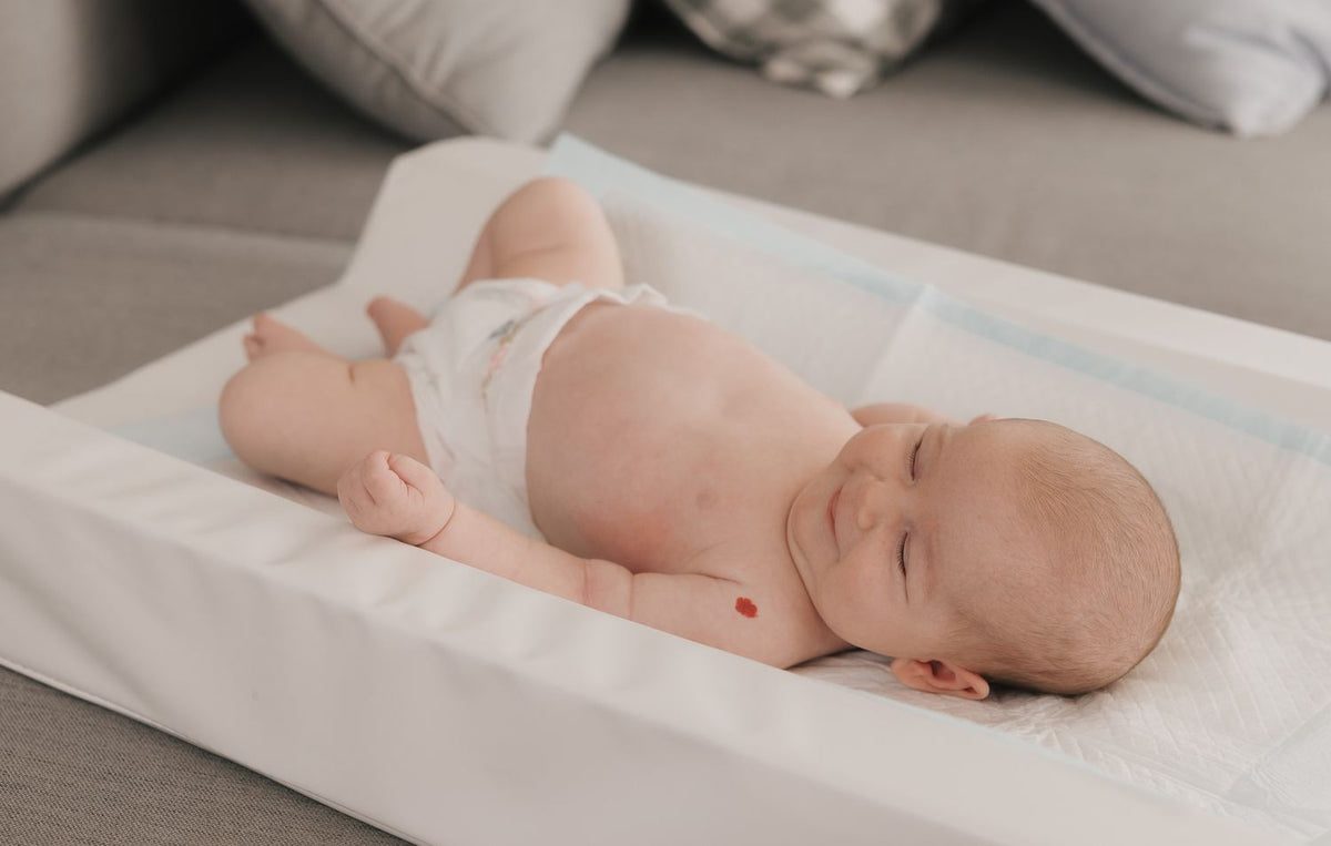 A newborn on a change mat in a correctly fitted nappy, the waistband sitting above the top of the bottom and the leg elastic down in the crease, with no gaping at either leg