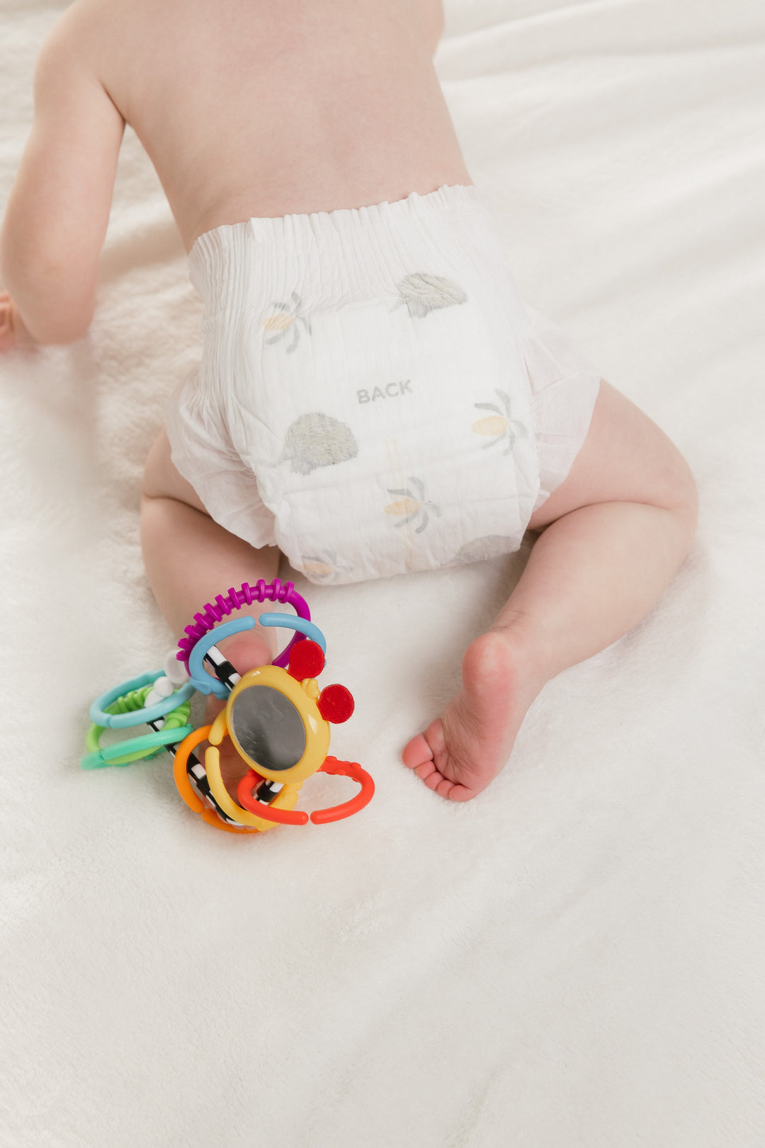 Baby on a white blanket in a Little Puggles nappy, seen from behind, with a toy beside them