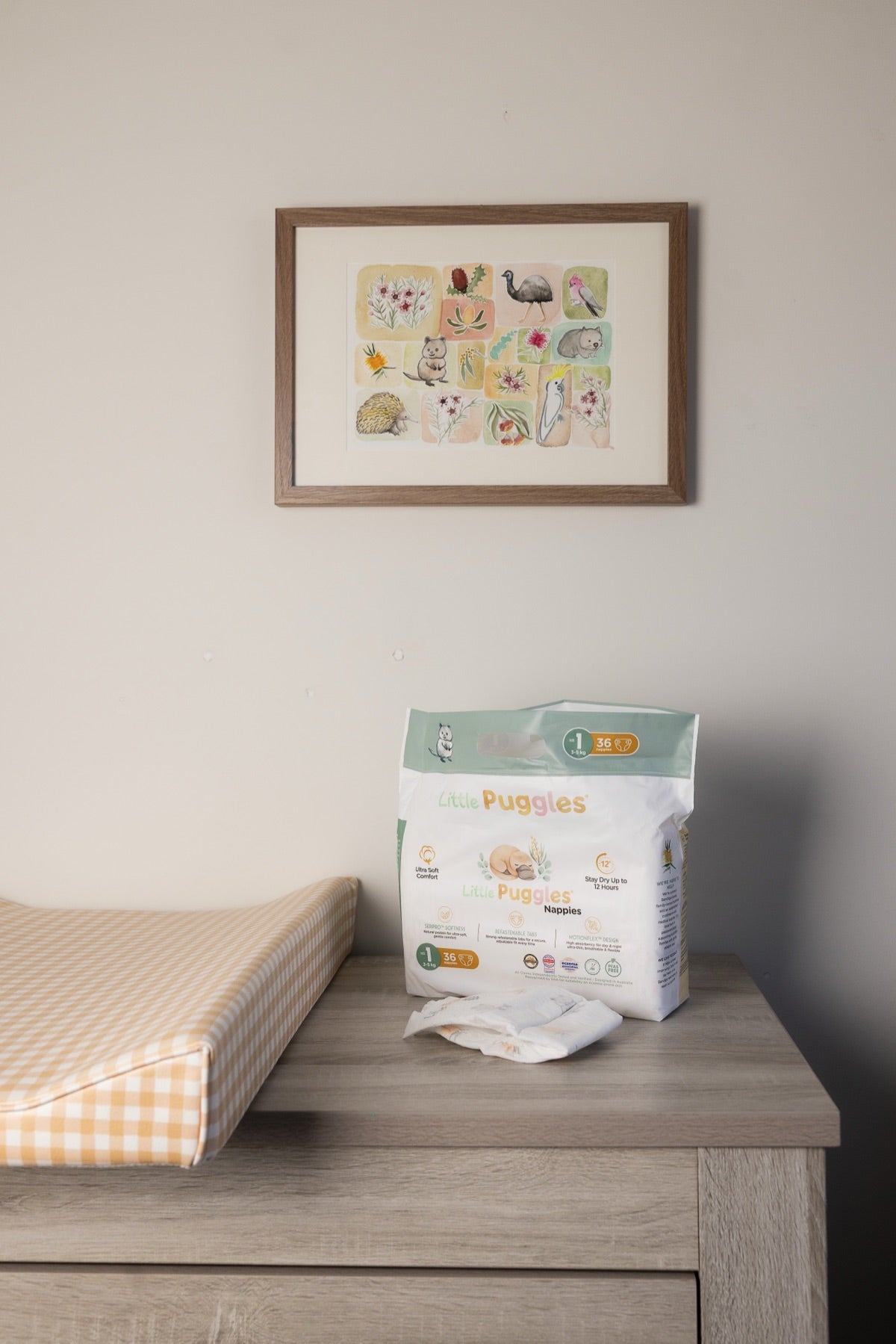 A pack of Little Puggles nappies standing on a nursery change table beside a flat quilted change mat, under a framed print of Australian animals and plants.