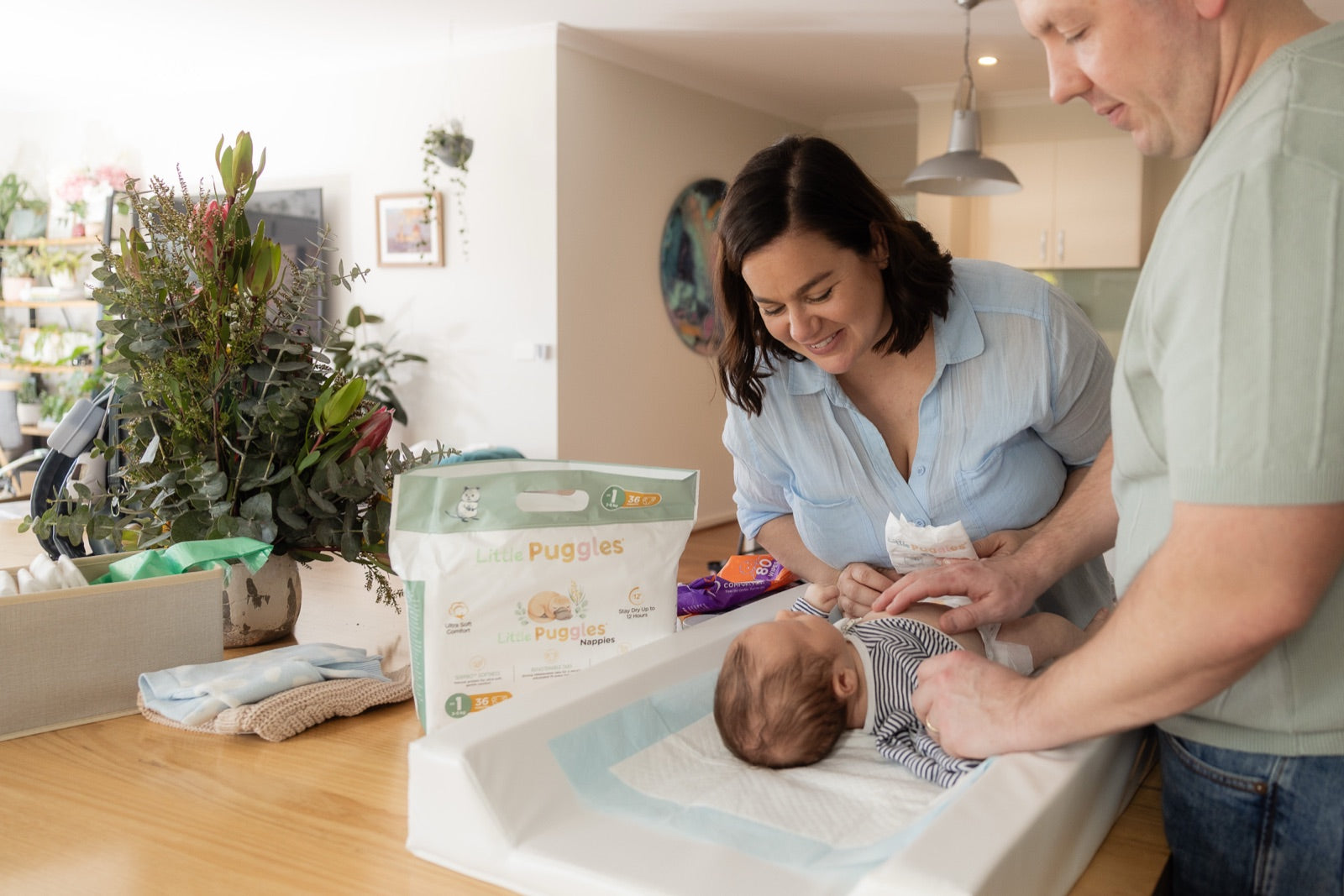 Two parents leaning over a change table in a kitchen, Louis on the mat between them, a sealed pack of Little Puggles nappies standing on the bench and a woven box of native foliage to the left of frame.