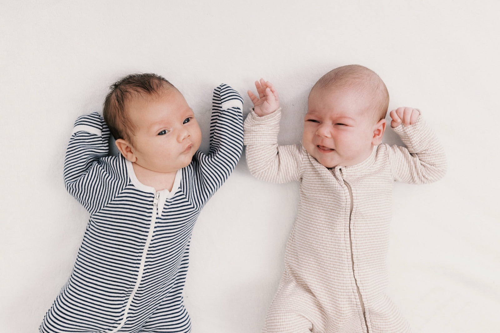 Two babies lying side by side on a cream blanket in zip sleepsuits, one in navy stripes and one in oatmeal, both with their arms up.
