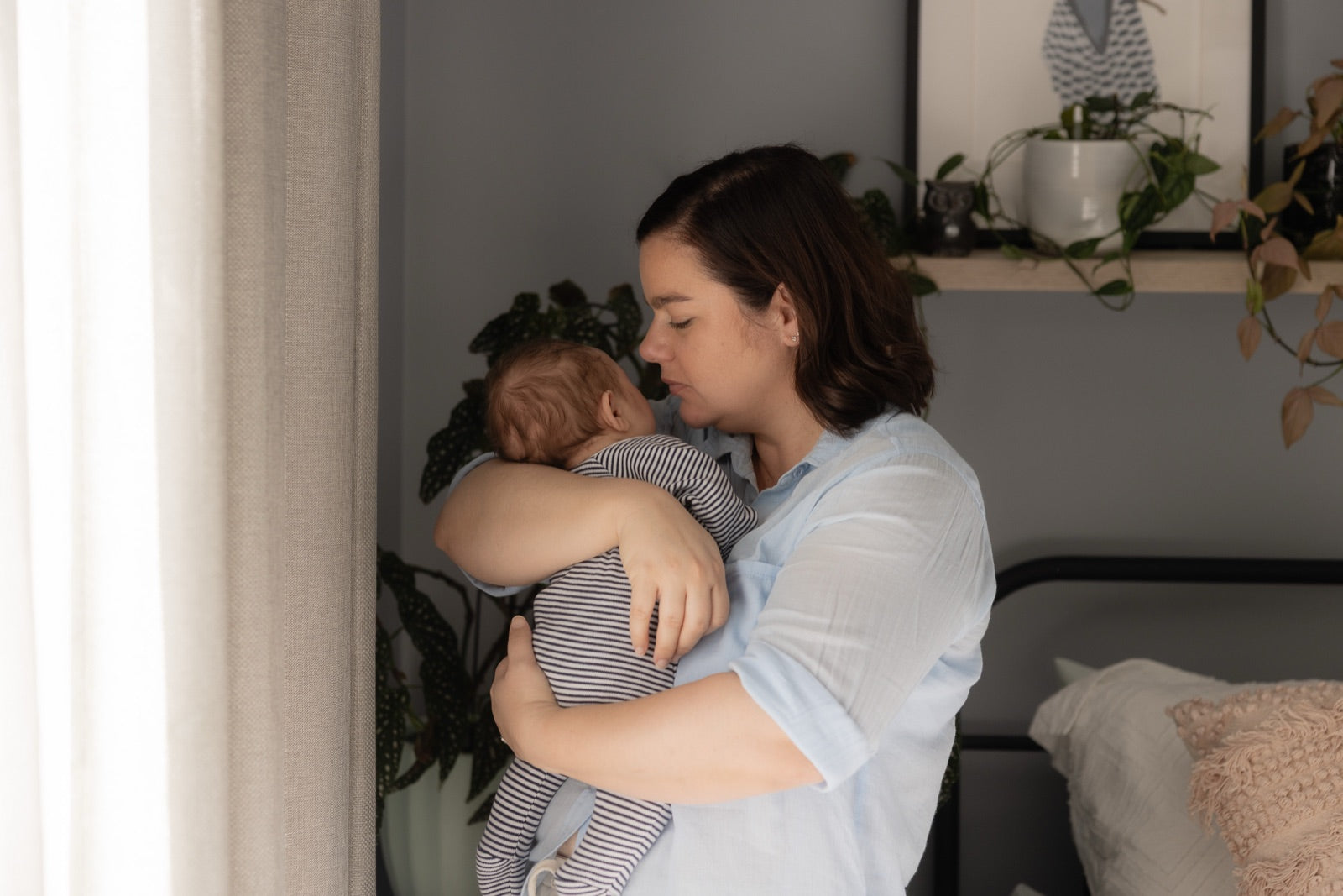 Emma stands at a bedroom window in soft daylight holding Louis against her shoulder, his head tucked under her chin, a shelf of trailing plants behind them.