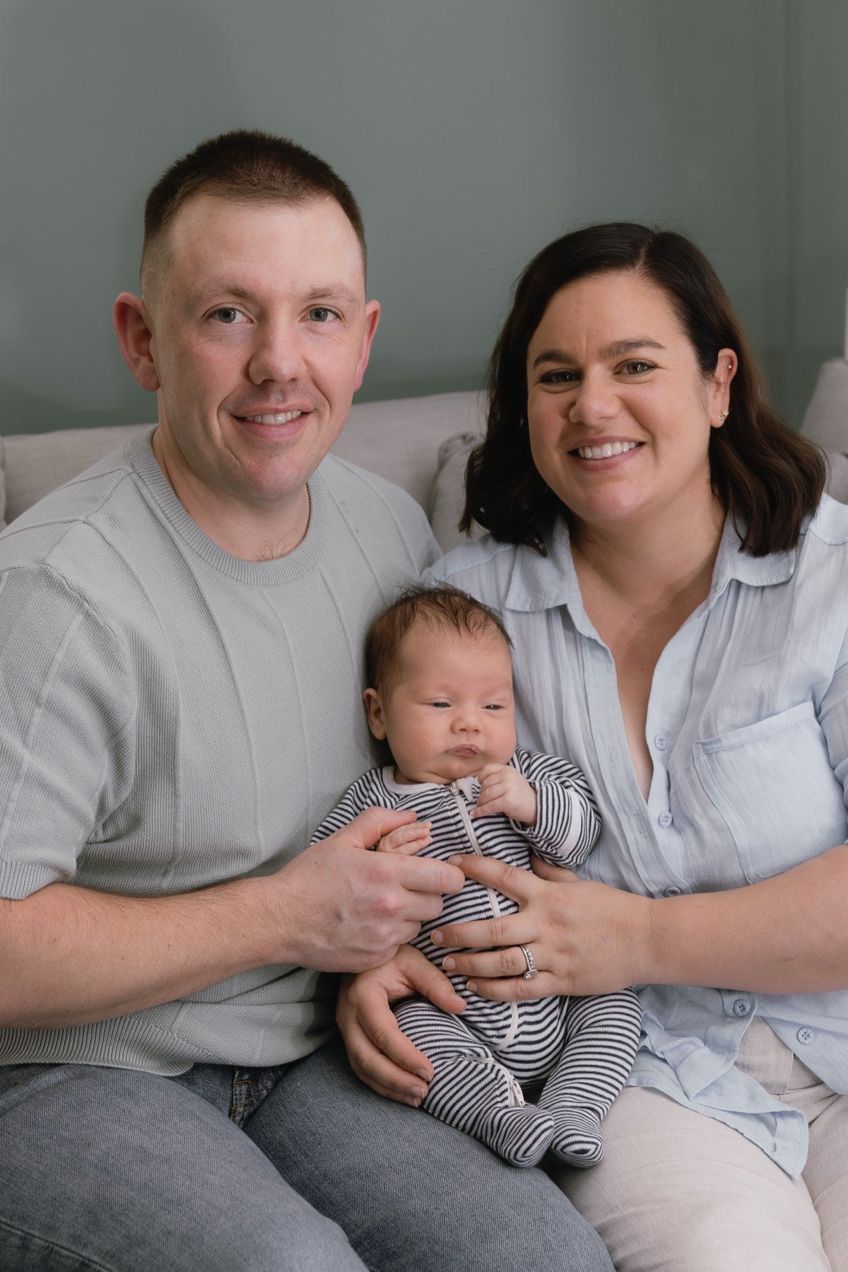 Little Puggles' two co-founders sitting together on a lounge, both smiling at the camera, with their baby son held between them.